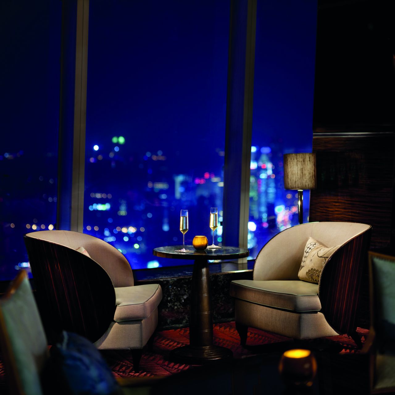 Two cocoon-like armchairs and a table with two glasses of champagne set next to large windows with evening city views