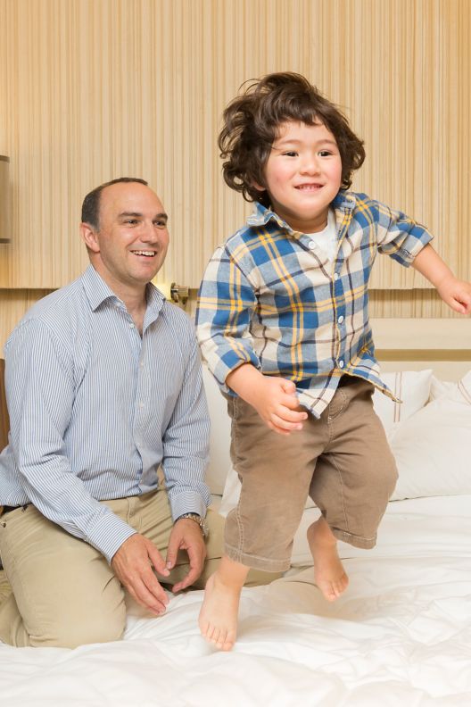 A dad smilingly sits on the bed and watches his toddler son jump on it.