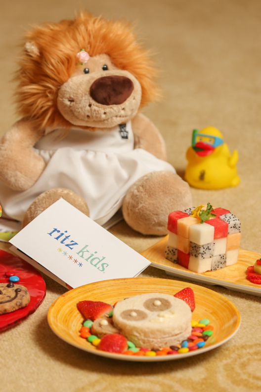 A stuffed lion sits on the floor surrounded by plates of treats, a bath toy and information about the Ritz Kids program