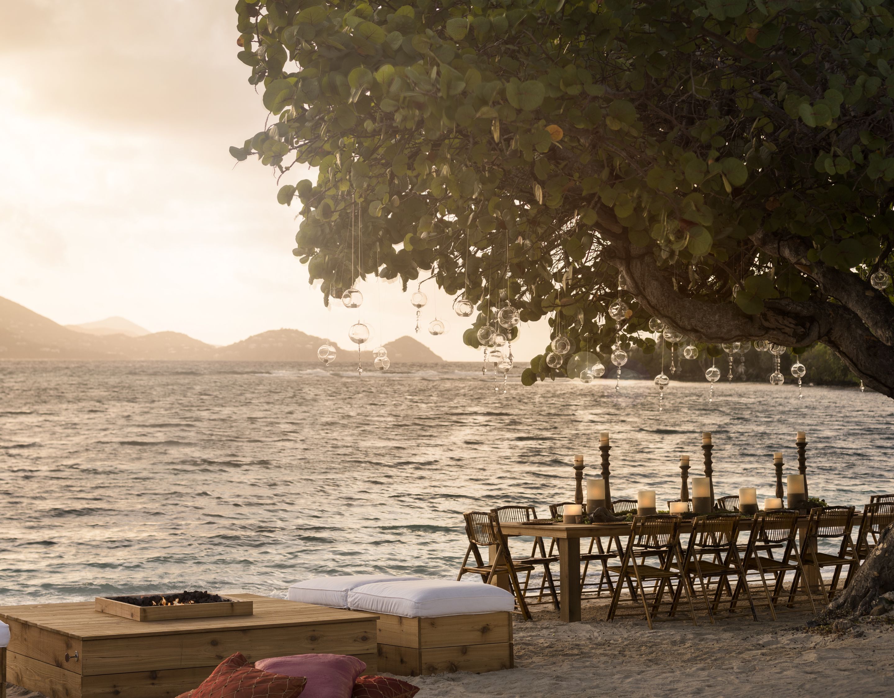 A long dining table and seating area set up near a large tree at the ocean?s edge