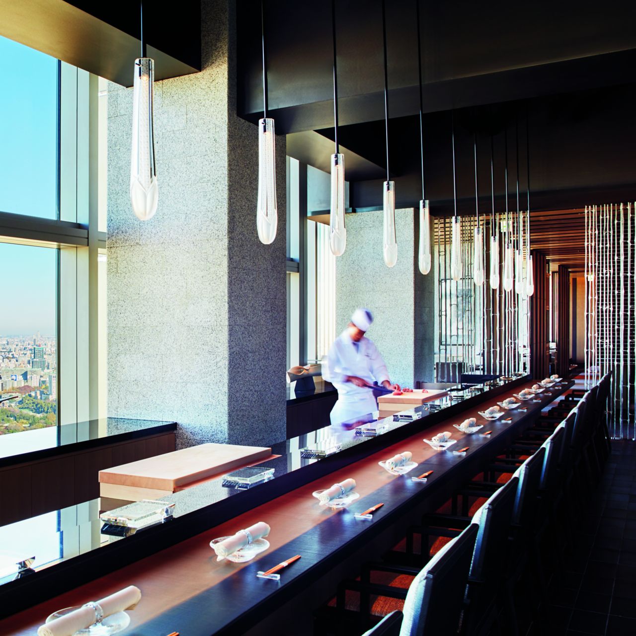 Tables lined up along floor-to-ceiling windows overlooking Tokyo in Hinokizaka with a chef working at the far end
