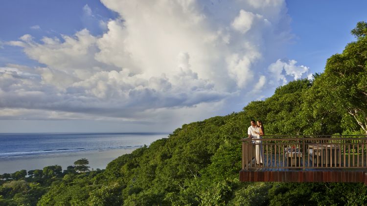 Ein Mann und eine Frau auf einem Holzbalkon mit Blick auf üppiges Grün und das Meer