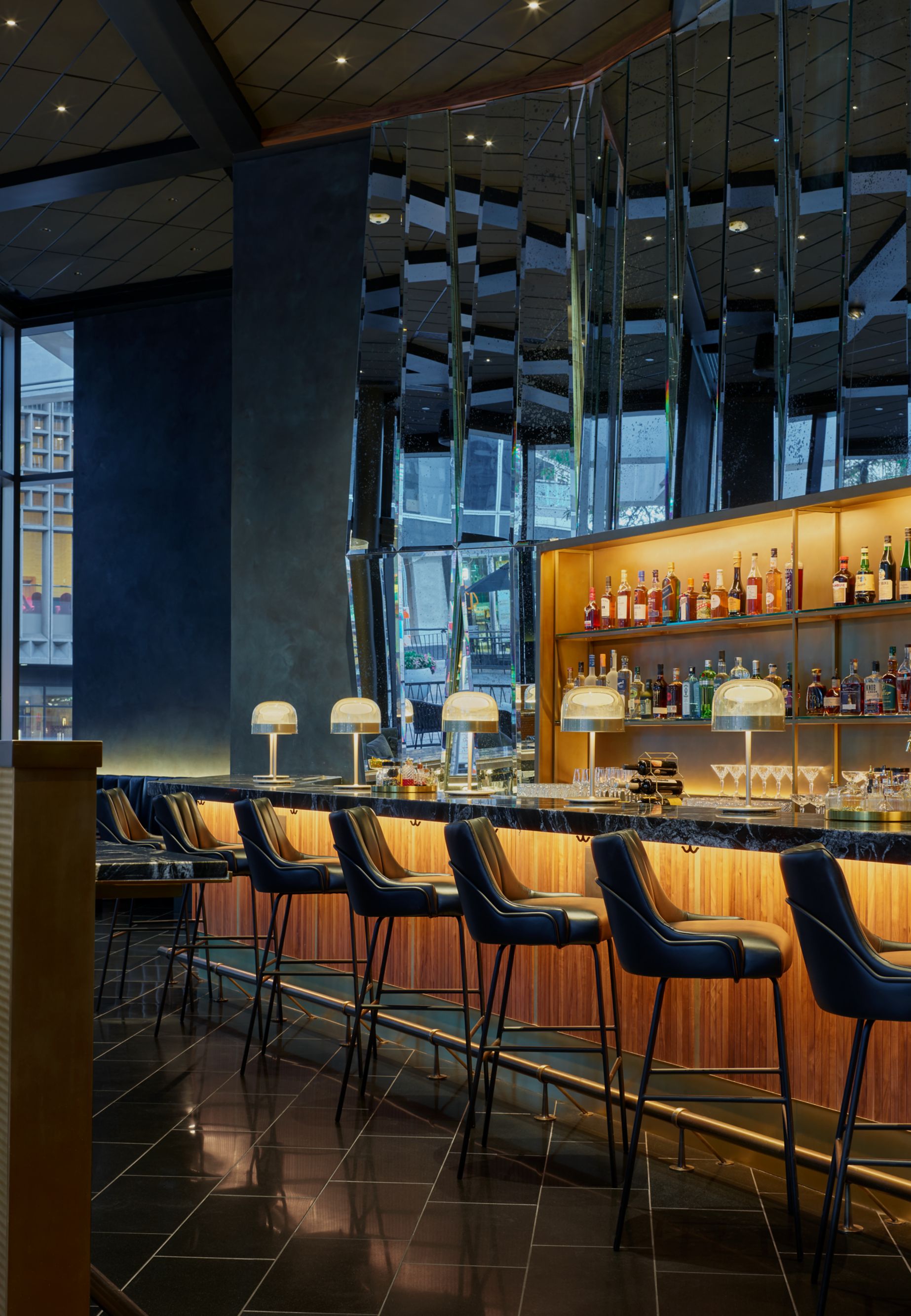A dimly lit bar with stools along the bar counter facing shelves of liquor bottles.