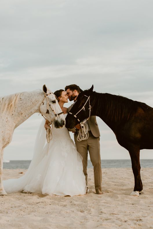 Bride and groom holding reins to horses on beach. 