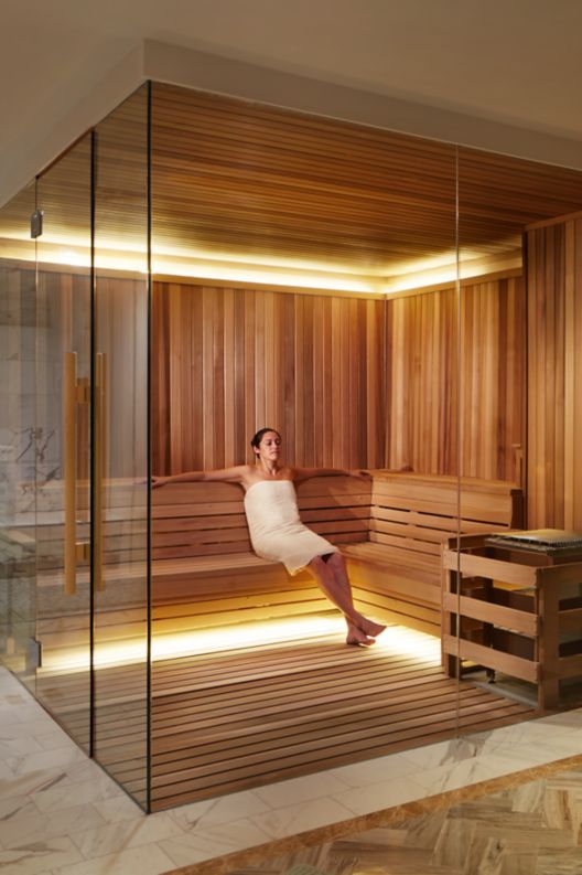 A woman in a towel relaxes in a large, glass-enclosed sauna