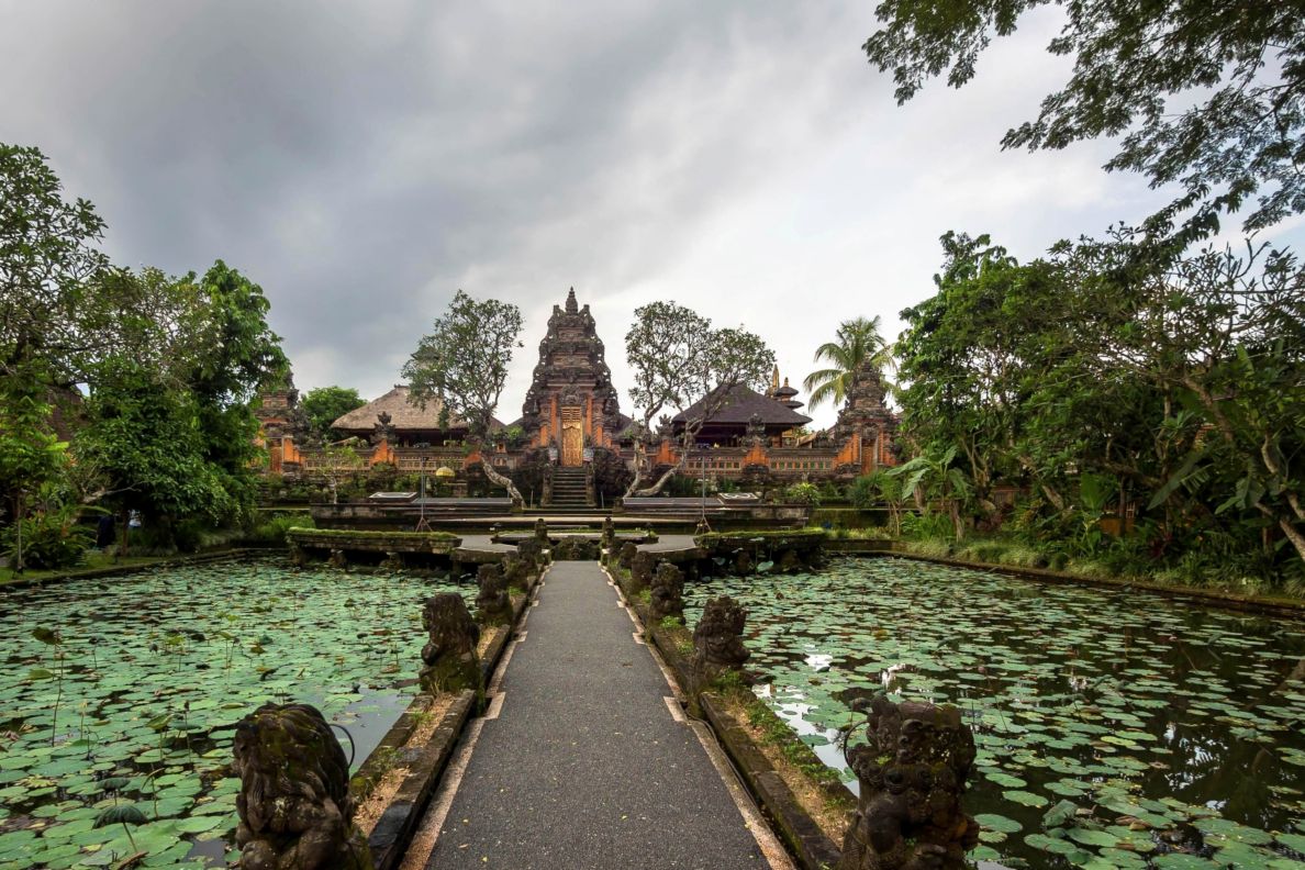 Long pathway flanked by water with lily pads leads to an orange temple under gray skies