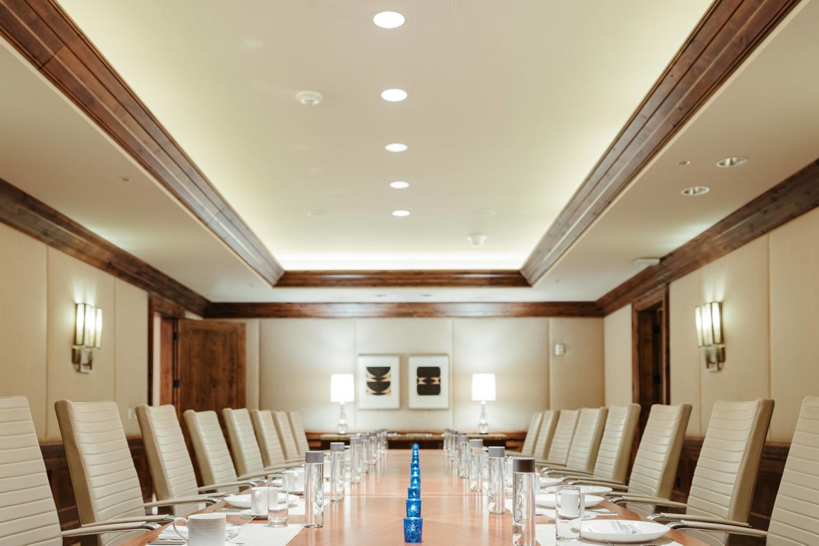 Meeting room with silverware on a rectangular table and tucked in arm chairs.