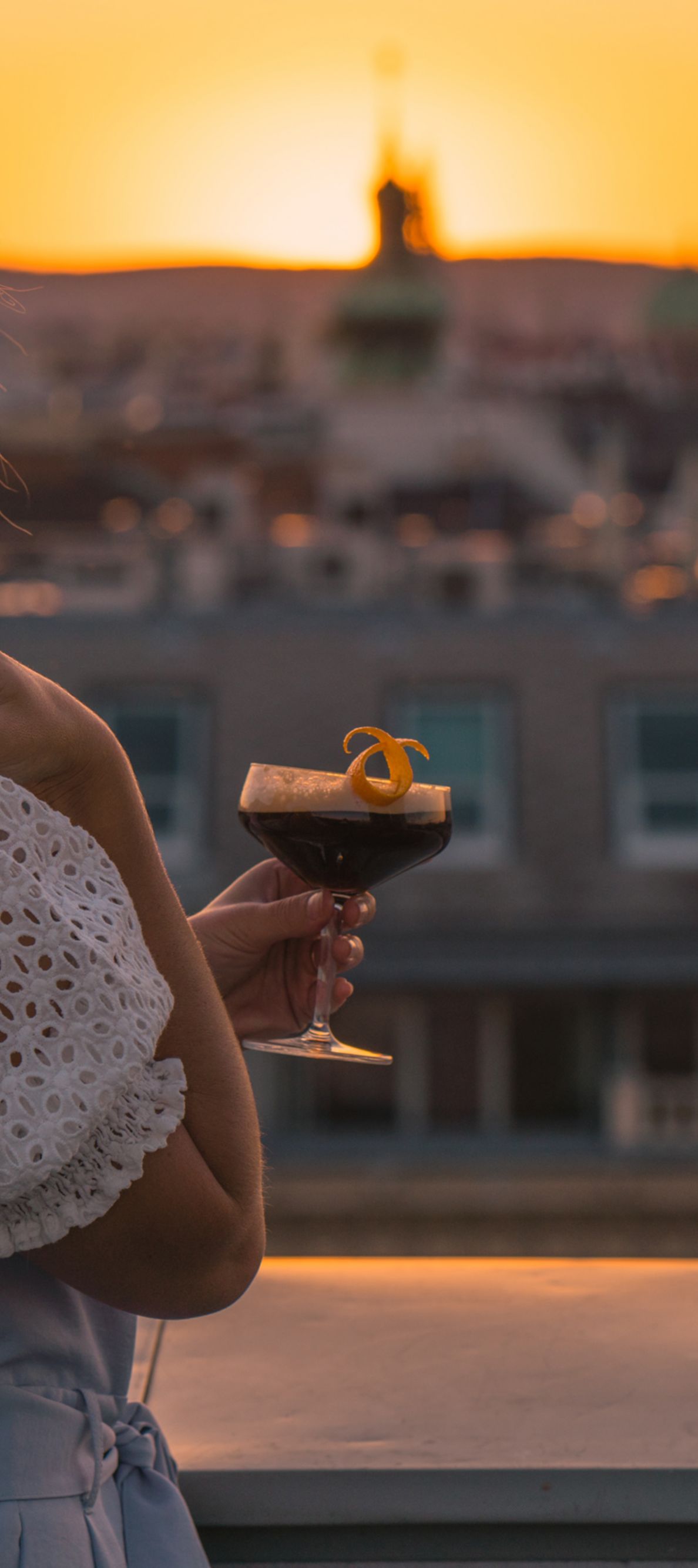 A lady looking over the rooftop at sun set holding a drink in hand. 
