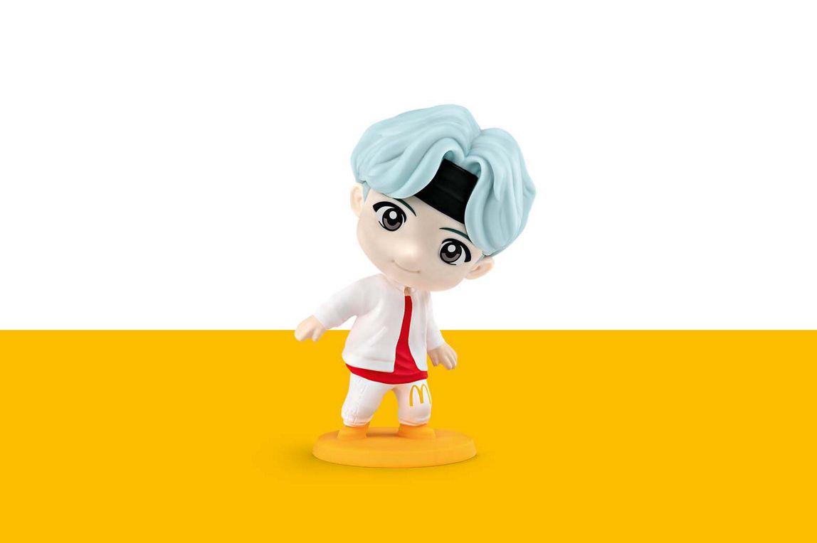 SUGA hat blaue Haare und trägt ein weißes Outfit mit einem goldenen M auf dem linken Hosenbein. 