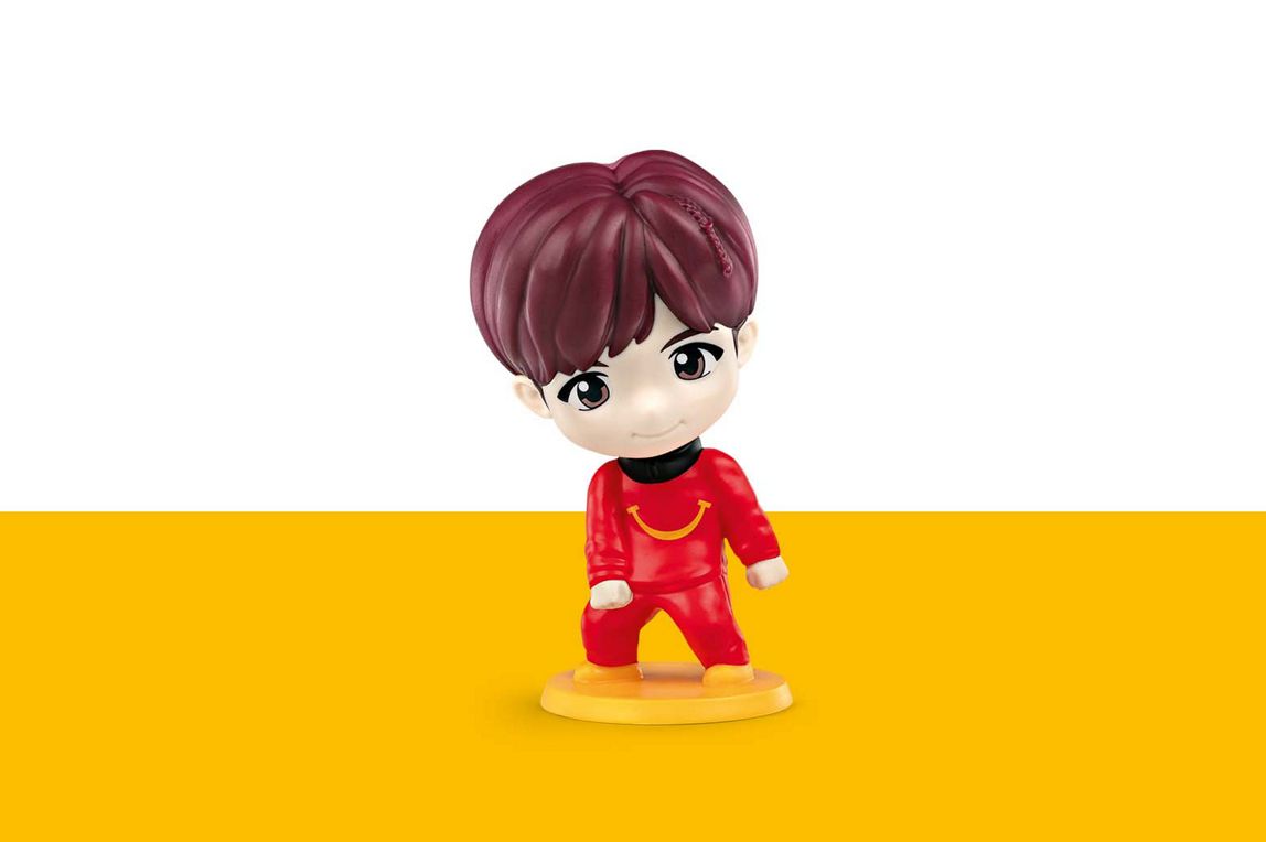j-hope hat braune Haare und trägt ein rotes Outfit mir einem gelb lächelndem Bund auf der Brust. 