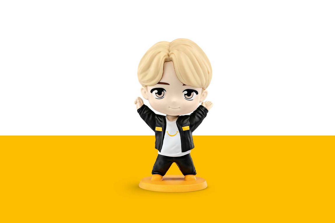 Jimin hat blonde Haare und trägt einen schwarzen Anzug und ein weißes T-Shirt mit einem gelben lächeln drauf. 