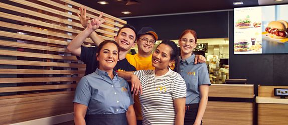 Fünf McDonald’s Crew-Mitglieder unterschiedlichen Alters und unterschiedlicher Herkunft stehen gemeinsam im Restaurant und lächeln in die Kamera.
