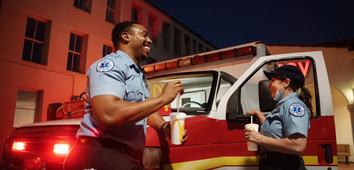 Two paramedics standing next to an ambulance smiling and eating McDonald's food and drinks