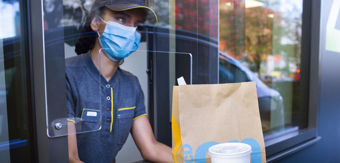 A McDonald's employee wearing a face mask holding a bag of food and a drink at the Drive Thru window