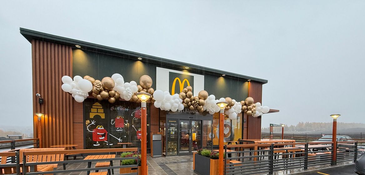 Utsiden av en McDonald's-restaurant med trepanel og store vinduer. Over inngangspartiet henger det dekorasjoner av hvite og gullfargede ballonger. Det er utemøbler i form av trebenker og bord, med rekkverk som omgir uteserveringsområdet. På vinduene er det klistremerker med McDonald's-karakterer, inkludert Happy Meal-figurer. Parkeringen foran restauranten er våt, noe som tyder på at det nylig har regnet, og i bakgrunnen kan man se et vinterlig landskap. Foto.