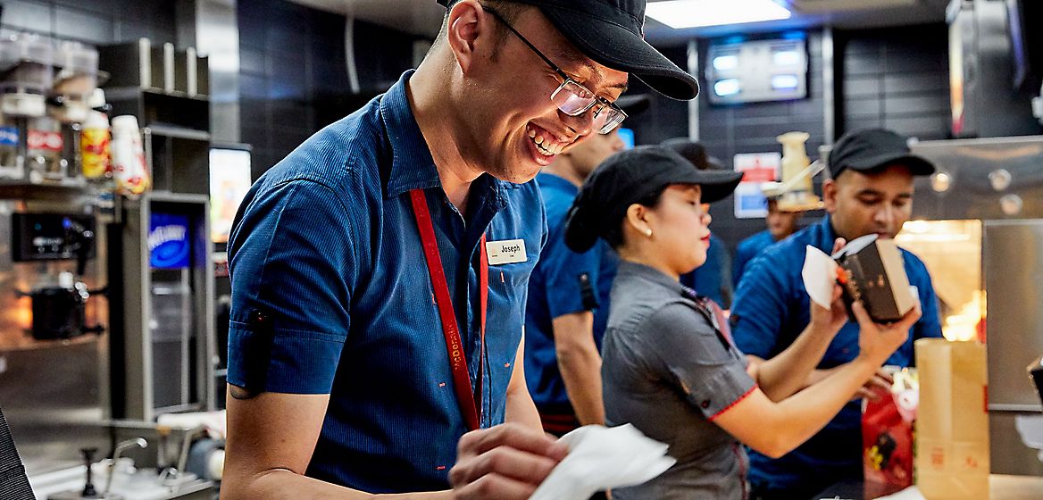 A McDonald's employee smiling in the foreground with other employees working in the background