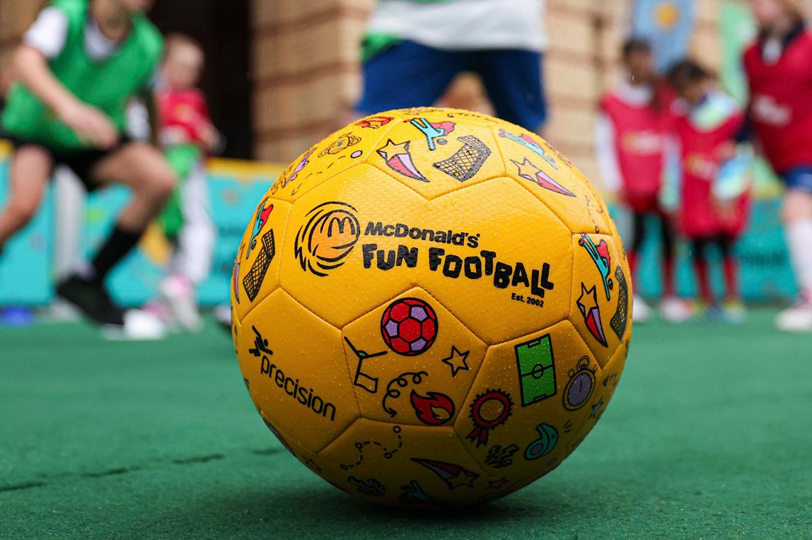 Close-up of a Fun Football football on a pitch with children playing behind it.