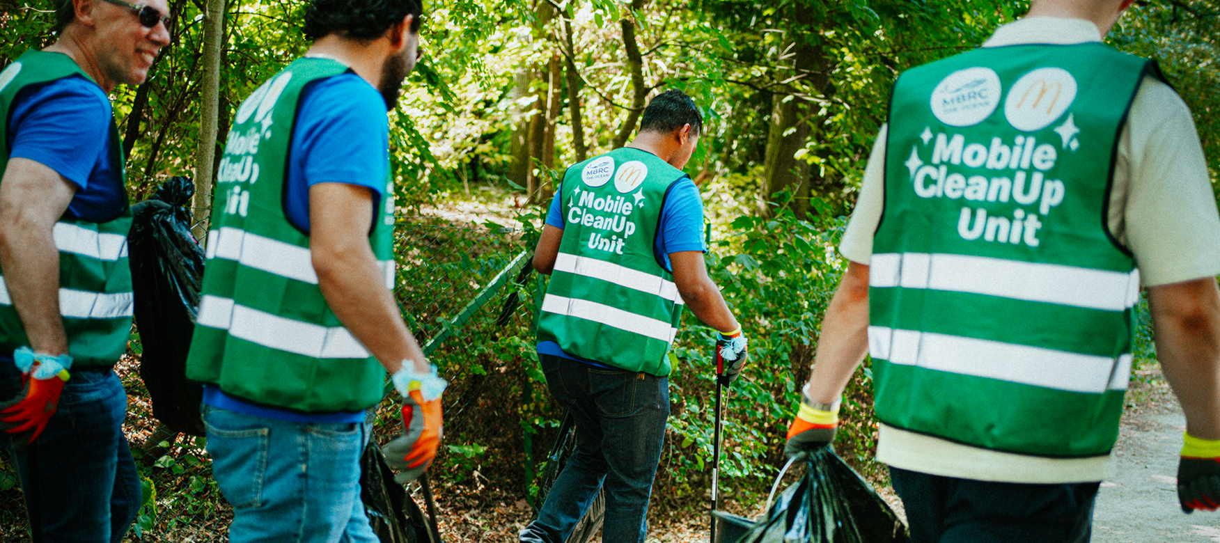 Vier Freiwillige in Warnwesten der Mobile CleanUp Unit sammeln Müll an einem Weg im Grünen