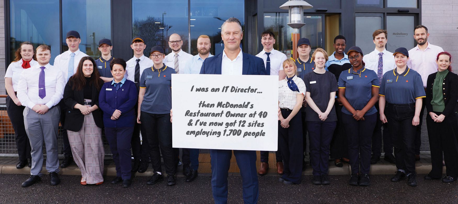 Multi-site franchisee Carel Venter with team outside a McDonald’s restaurant holding a sign: ‘I was an IT Director… then McDonald’s Restaurant Owner at 40 & I’ve now got 12 sites employing 1,700 people’.