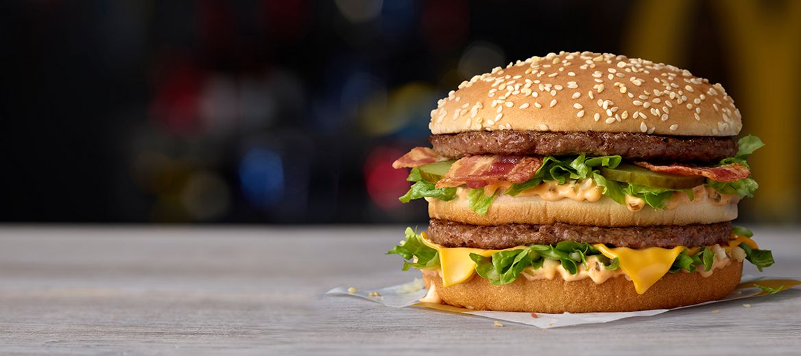 Two charred beef patties, crispy bacon, a slice of melted cheese, lettuce, onion, pickles, and orange Big Mac™ sauce in a sesame topped bun.