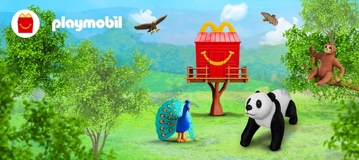 A red Happy Meal box treehouse sits in a field, with mountains in the backdrop and trees in the foreground. Playmobil eagles fly in the blue sky, a Playmobil sloth sits in a tree and a peacock and panda stand in front of the treehouse on the grass. 