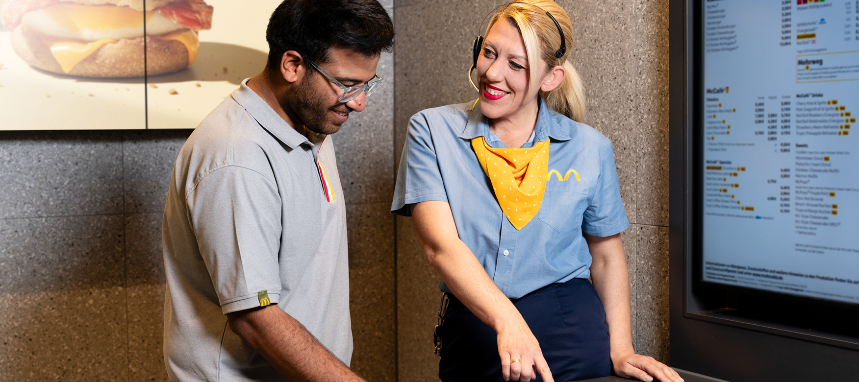 Zwei McDonald’s Crew-Mitglieder arbeiten gemeinsam an einem Bestellterminal im Restaurant und lächeln.