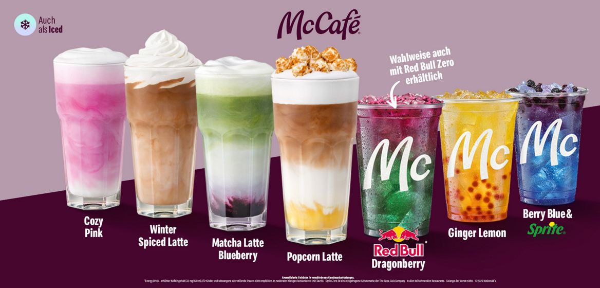 Werbegrafik von McCafé mit sieben bunten Herbstgetränken vor einem zweifarbigen Hintergrund in Altrosa und Dunkelviolett. Von links nach rechts sind Cozy Pink, Winter Spiced Latte, Matcha Latte Blueberry und Popcorn Latte in hohen Gläsern mit cremigen Schichten und Sahnehauben zu sehen. Daneben stehen drei erfrischende Kaltgetränke mit Eiswürfeln: Red Bull Dragonberry in Pink-Grün, Ginger Lemon in Gelb-Orange und Berry Blue & Sprite in Blau-Violett. Oben befindet sich das McCafé-Logo sowie der Hinweis, dass die Getränke auch als Iced-Varianten beziehungsweise teilweise mit Red Bull Zero erhältlich sind.