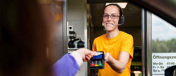 McDonald’s-Mitarbeiterin mit Headset reicht einem Gast am Drive-in-Schalter ein Kartenlesegerät.