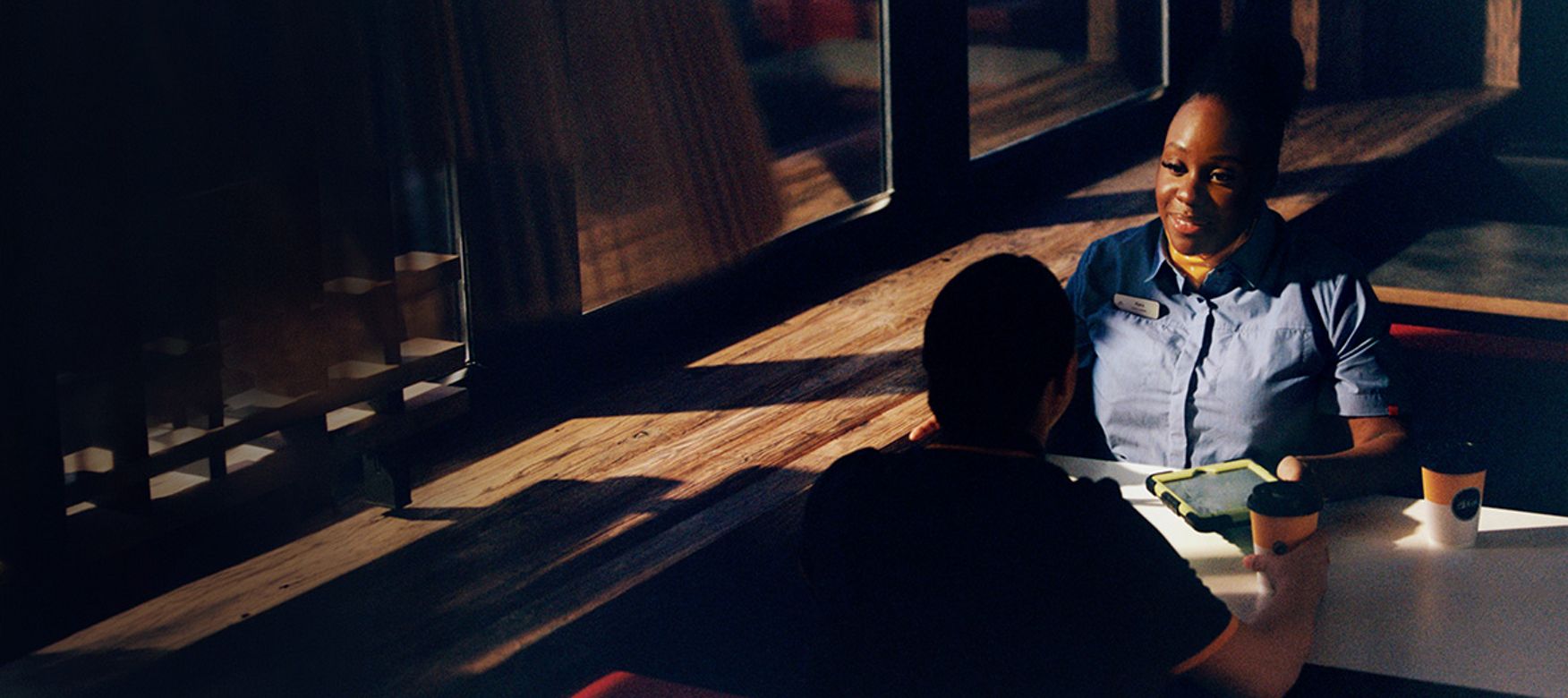 Two McDonald’s staff sitting at a table in a McDonald’s restaurant. Two takeaway coffee cups and a tablet or menu device are placed on the table between them. Sunlight comes through large windows, casting shadows across the seating area.