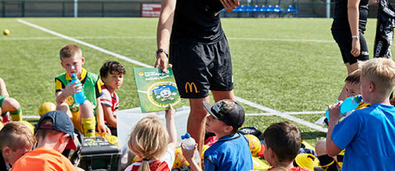 Group of young kids sitting around a football coach handing out fun football activity book