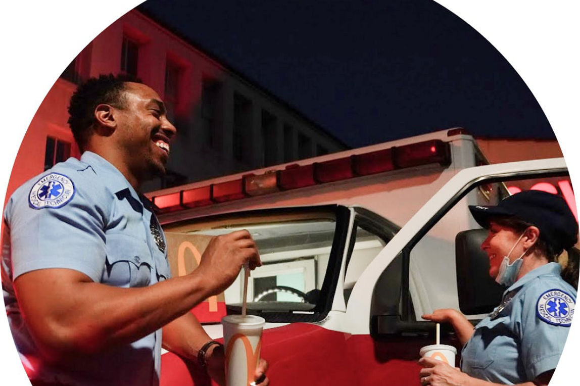 Two paramedics standing next to an ambulance smiling and eating McDonald's food and drinks