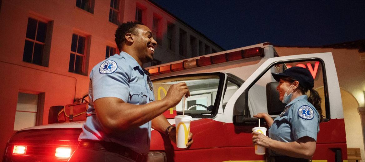 Two paramedics standing next to an ambulance smiling and eating McDonald's food and drinks