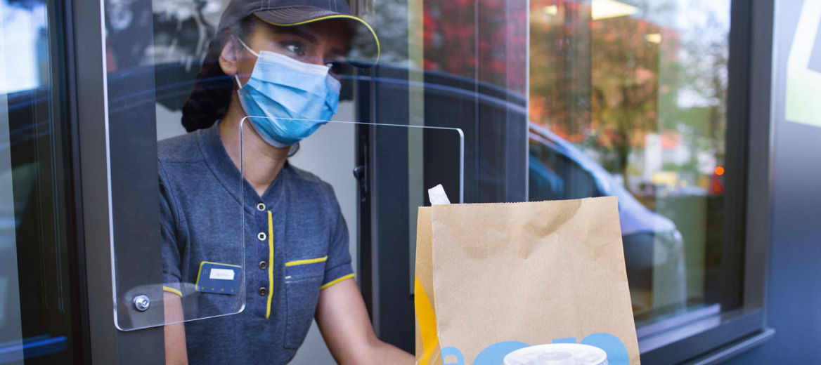 A McDonald's employee wearing a face mask holding a bag of food and a drink at the Drive Thru window