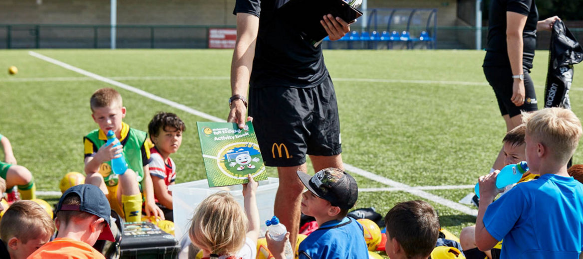 Group of young kids sitting around a football coach handing out fun football activity book