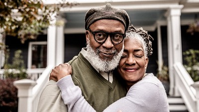 An old man with a beard is embraced by his wife