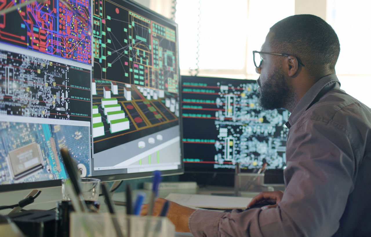 Stock image of an Afro-Caribbean male designing electronic circuit boards ( PCBs).
Heâ  s sitting at a desk with a large computer screen displaying a variety of apps & PCB illustrations.