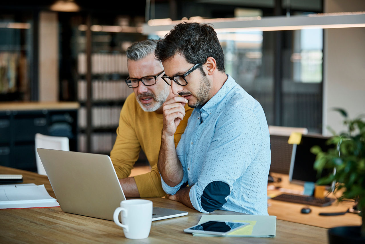 Businessmen using laptop together at desk. Male executives are working at textile factory. Professionals are in factory.