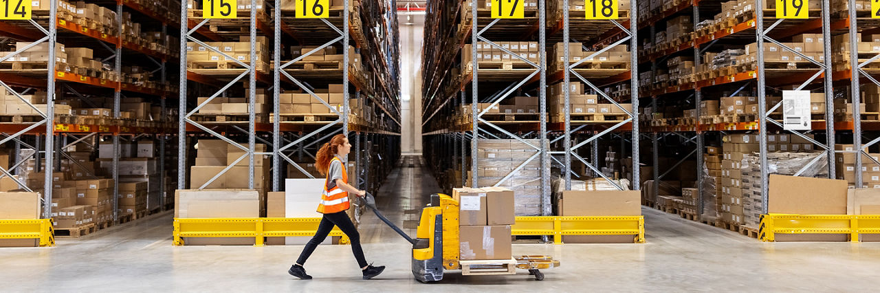 Side view of a female worker pushing hand truck with boxes in a storehouse. Warehouse employee walking with cardboard boxes on pallet jack along large racks.