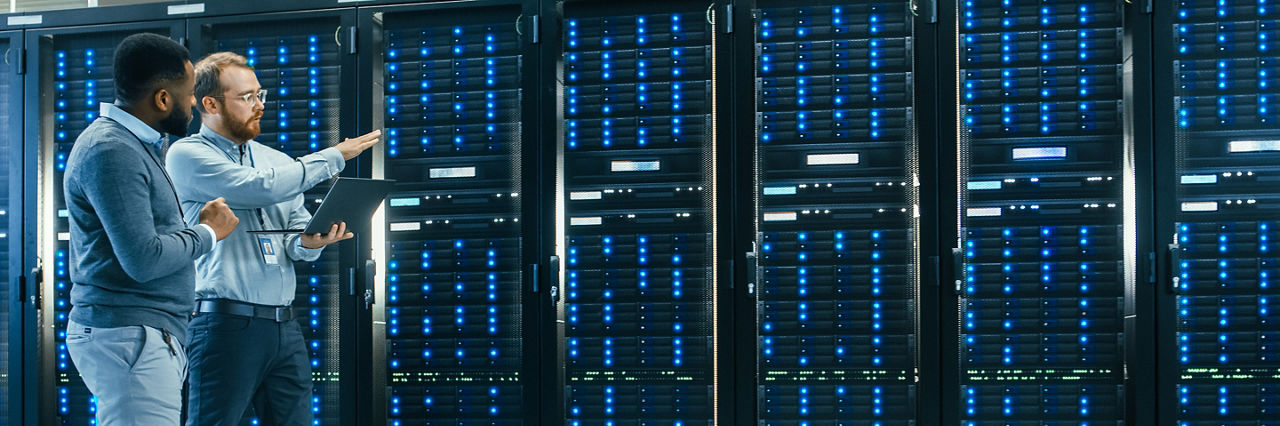 IT Technician with a Laptop Computer and Black Male Engineer Colleague are Talking in Data Center while Walking Next to Server Racks. Running Diagnostics or Doing Maintenance Work.