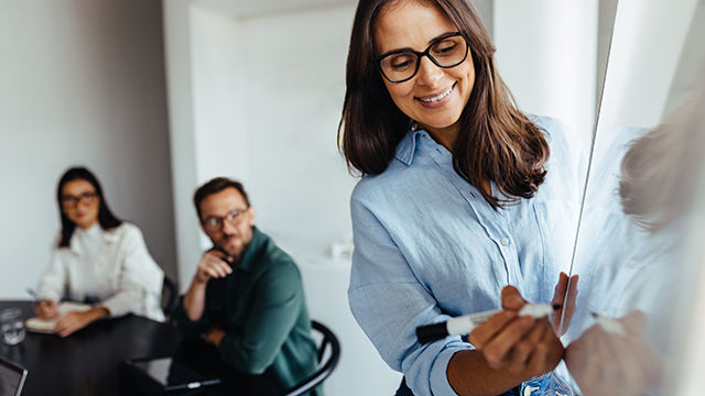 Business woman writing on a board during a meeting with her team in an office. Creative business woman giving a presentation of her ideas on a new project.