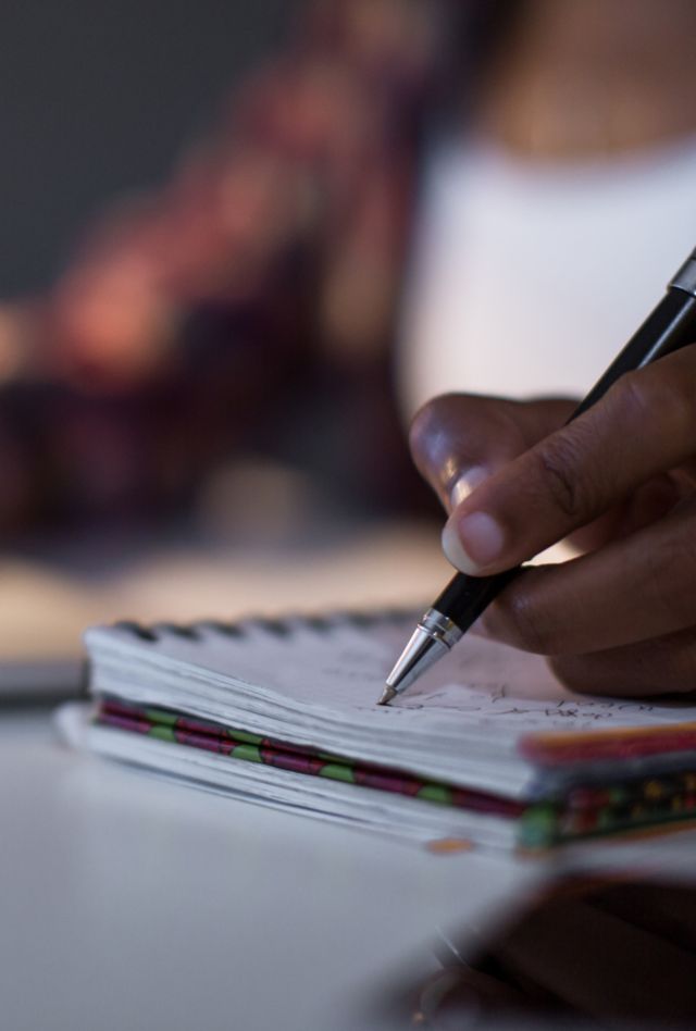 A student sits in front of a laptop writing in a notebook.