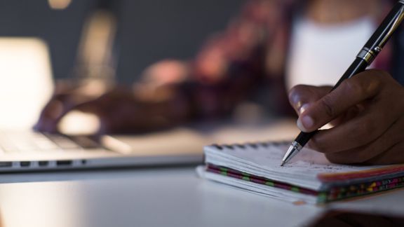 A student sits in front of a laptop writing in a notebook.