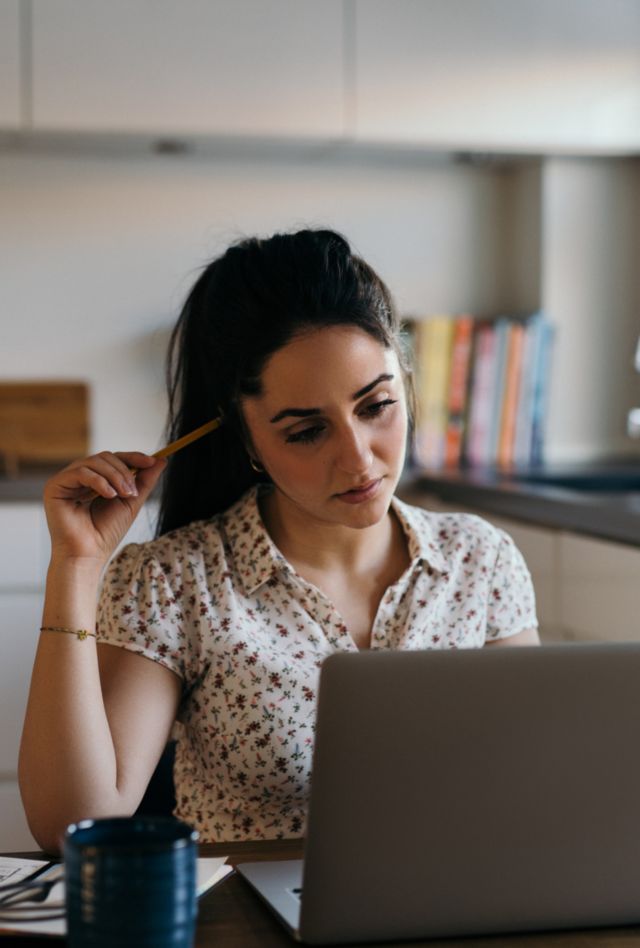 Young brunette woman sitting at table in kitchen and looking at laptop with pencil to her hairs, thinking while working or studying from home