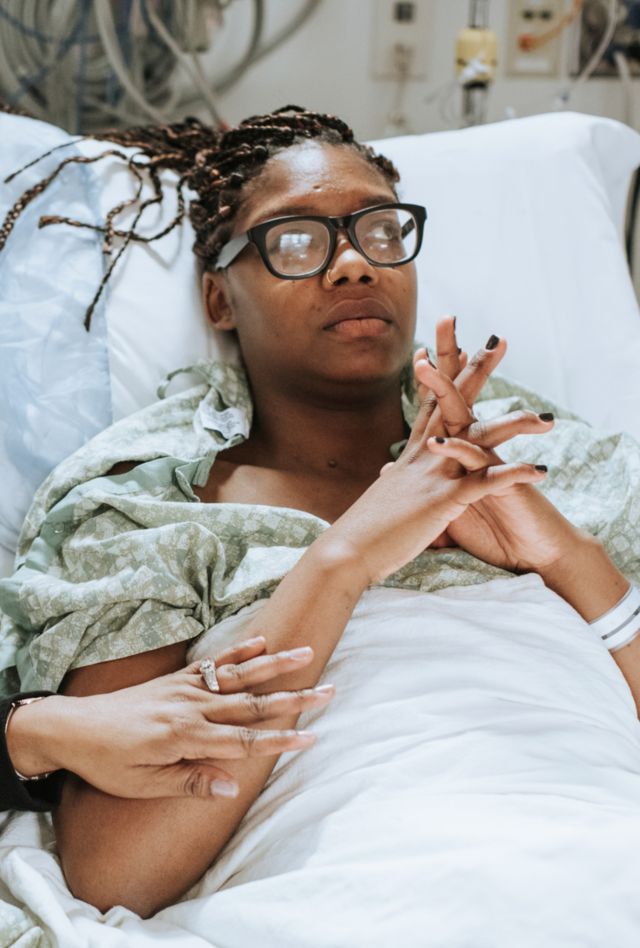 Attractive, young woman resting in bed of medical facility with fingers crossed, looking off into the distance as she anxiously awaits procedure. Her mother is seated at her side, gently rubbing her forearm to provide comfort.
