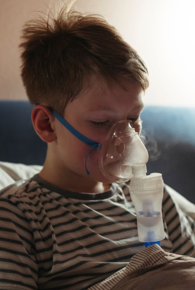 A little sick boy takes an inhalation while lying in bed in the evening.