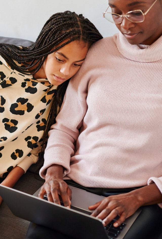Young girl sitting with her mother on their living room sofa and watching something on a laptop