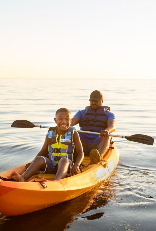 Young Black teen boy in front of tandem kayak grins widely as his father rests from rowing.  Golden hour light.