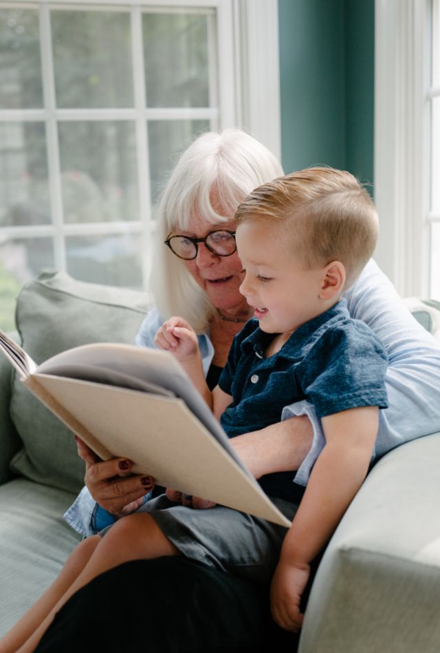 Grandparent sitting in a big chair with her grandson, looking through a book