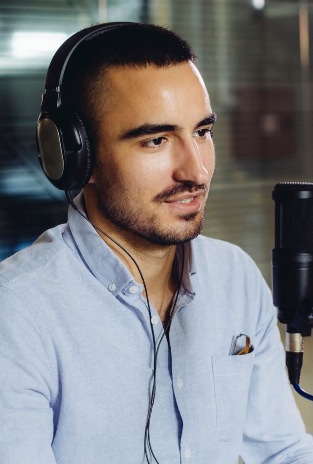 Young male entrepreneur recording a podcast in the office. He is focused and cheerful, while talking into the microphone. He is casually dressed in light blue shirt. 