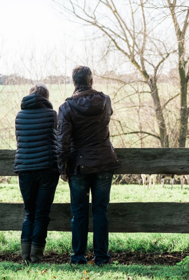 Two people stand in front of a wooden fence looking at a rural landscape